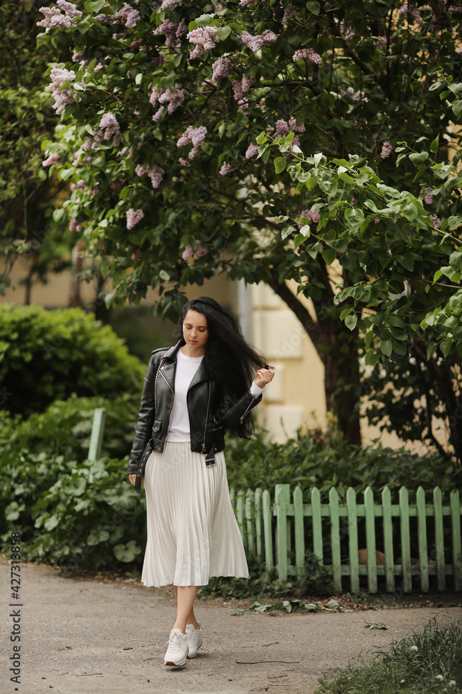 Pretty model girl in summer midi skirt and leather jacket posing near a bloomy tree. Young brunette woman in spring outfit posing outdoors Pretty model girl in summer midi skirt and leather jacket posing near a bloomy tree. Young brunette woman in spring outfit posing outdoors