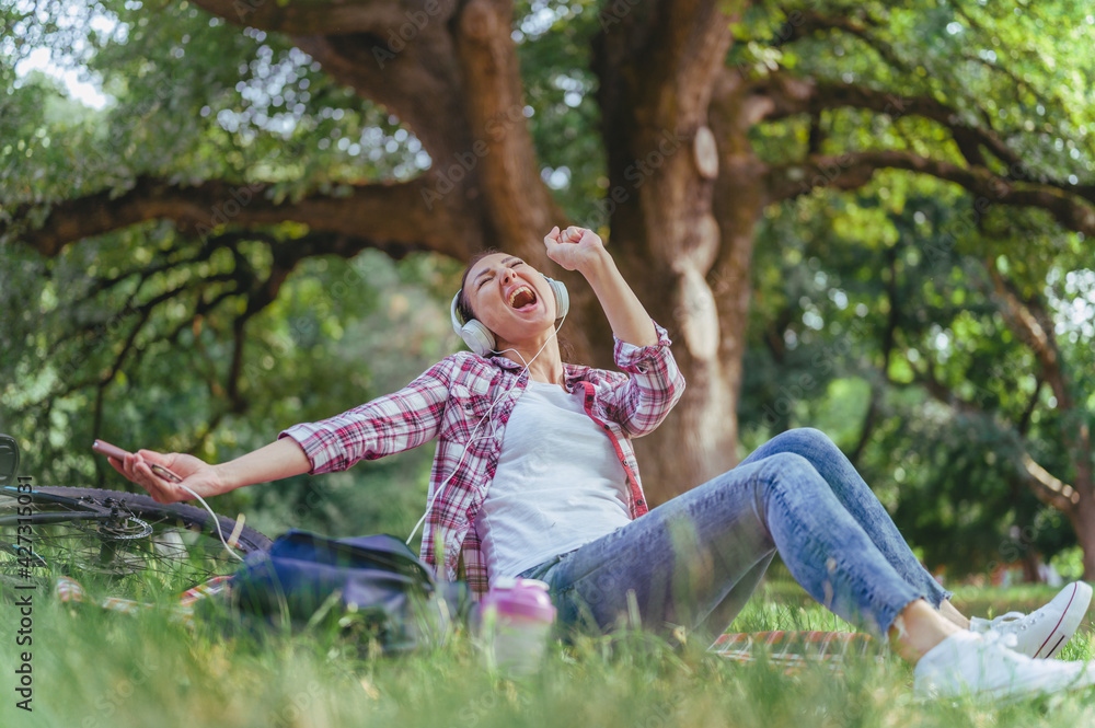 Woman with headphones sitting on a grass in a public park listening music over smartphone singing loud. Positive emotions Woman with headphones sitting on a grass in a public park listening music over smartphone singing loud. Positive emotions