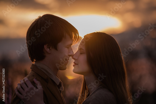 Silhouette of a happy couple in love at sunset in blooming peach rose gardens. Man stands in front of young woman, hugs, kisses with tenderness and passion. Close-up portrait of lovers. Romantic date