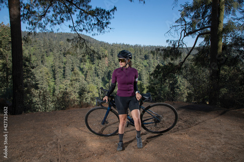 Joven mujer ciclista posando con su bicicleta en un mirador con un paisaje boscoso al fondo. Ciclista profesional con casco y gafas de sol.