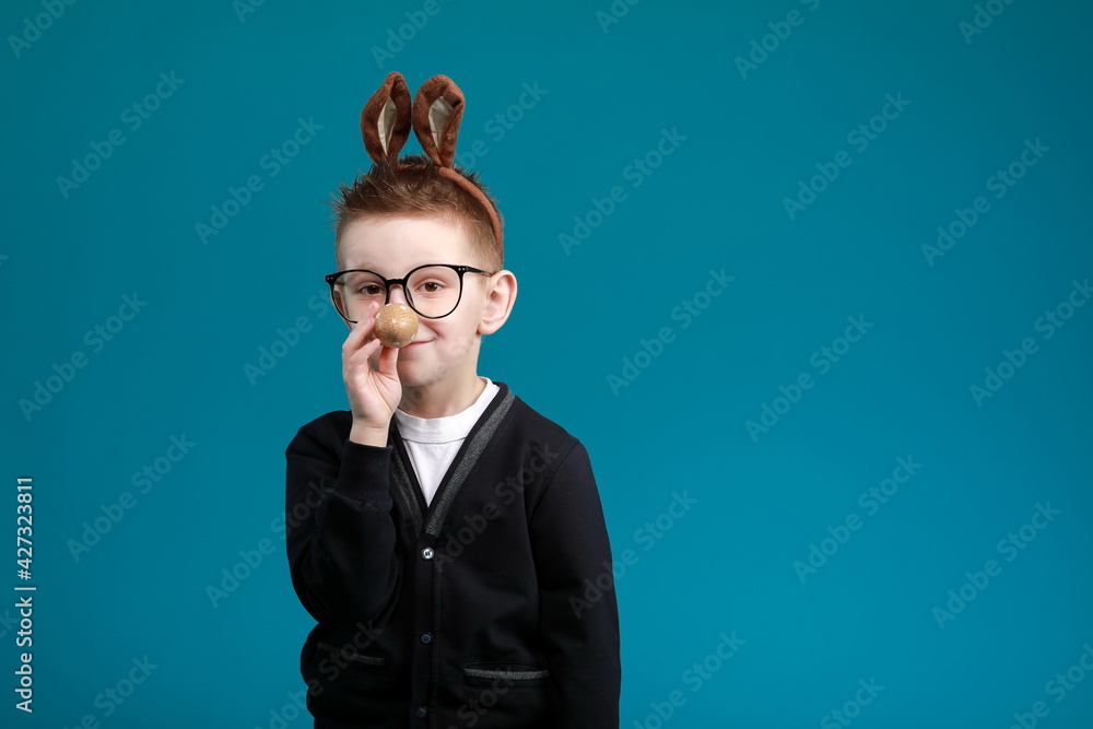 Cheerful boy in rabbit bunny ears on head on blue studio background. Excited crazy smiling happy child in glasses. Easter blue color of the year painting eggs in hands. Easter and holidays.