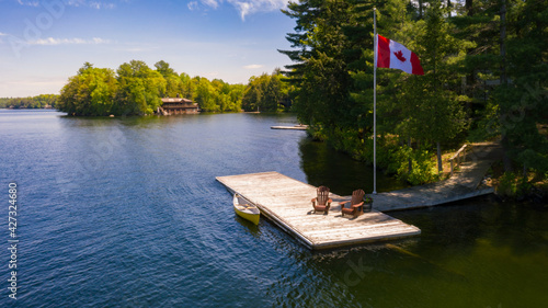 Fototapeta Naklejka Na Ścianę i Meble -  Aerial view of two Adirondack chairs on a wooden dock facing a lake in Muskoka, Ontario Canada. A yellow canoe is tied to the pier where a Canadian flag is flying. A cottage is visible in background.
