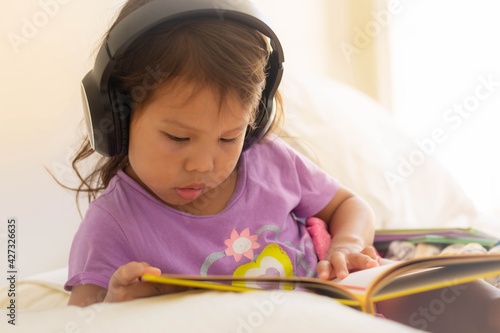 A little girl reading a book at home and listening to music wearing headphones. Audio books.