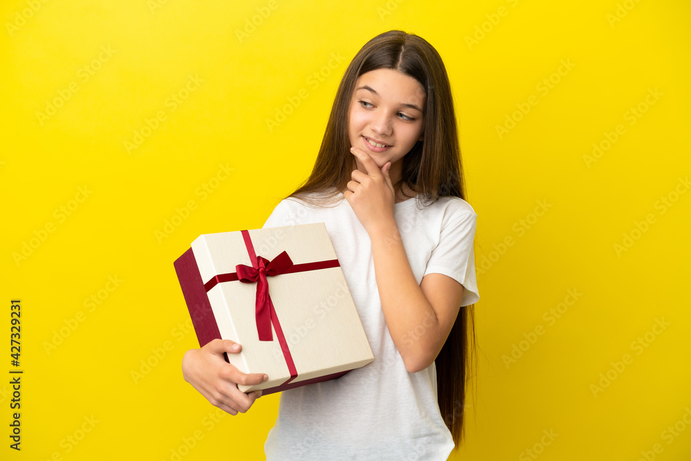 Little girl holding a gift over isolated yellow background looking to the side and smiling Little girl holding a gift over isolated yellow background looking to the side and smiling