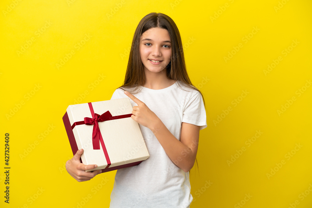 Little girl holding a gift over isolated yellow background pointing to the side to present a product Little girl holding a gift over isolated yellow background pointing to the side to present a product