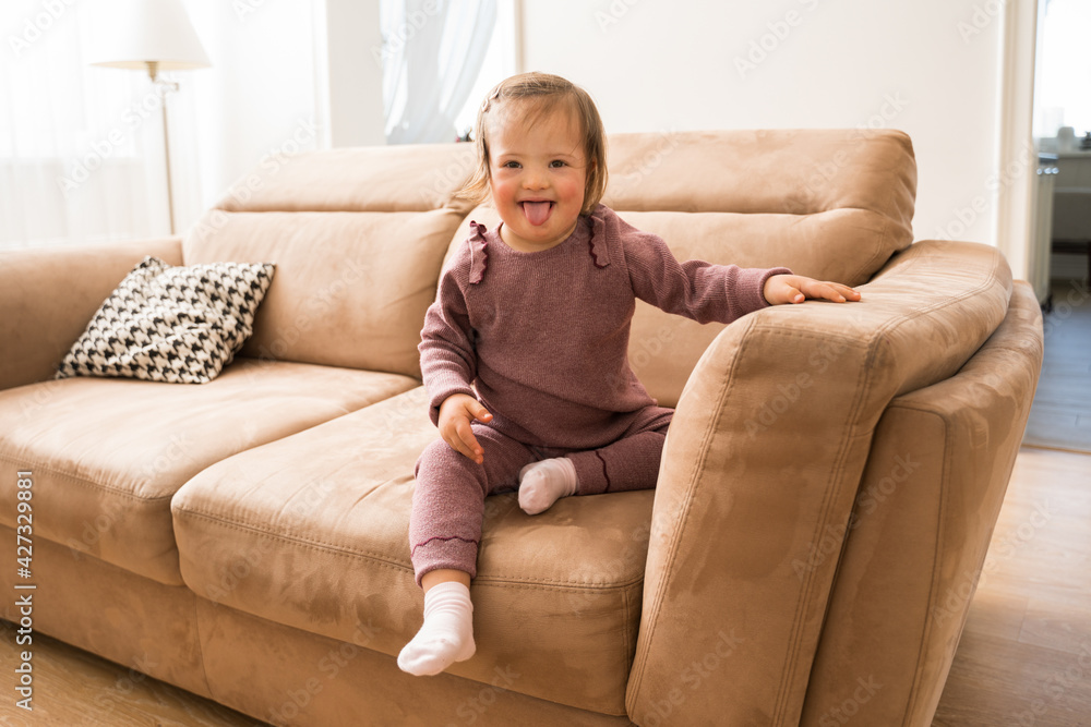 Girl with down syndrome sitting at the sofa and showing her tongue Girl with down syndrome sitting at the sofa and showing her tongue