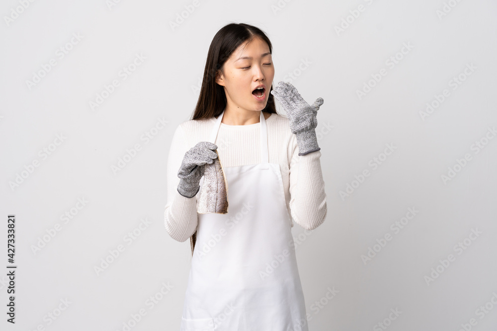 Chinese fishmonger wearing an apron and holding a raw fish over isolated white background yawning and covering wide open mouth with hand Chinese fishmonger wearing an apron and holding a raw fish over isolated white background yawning and covering wide open mouth with hand