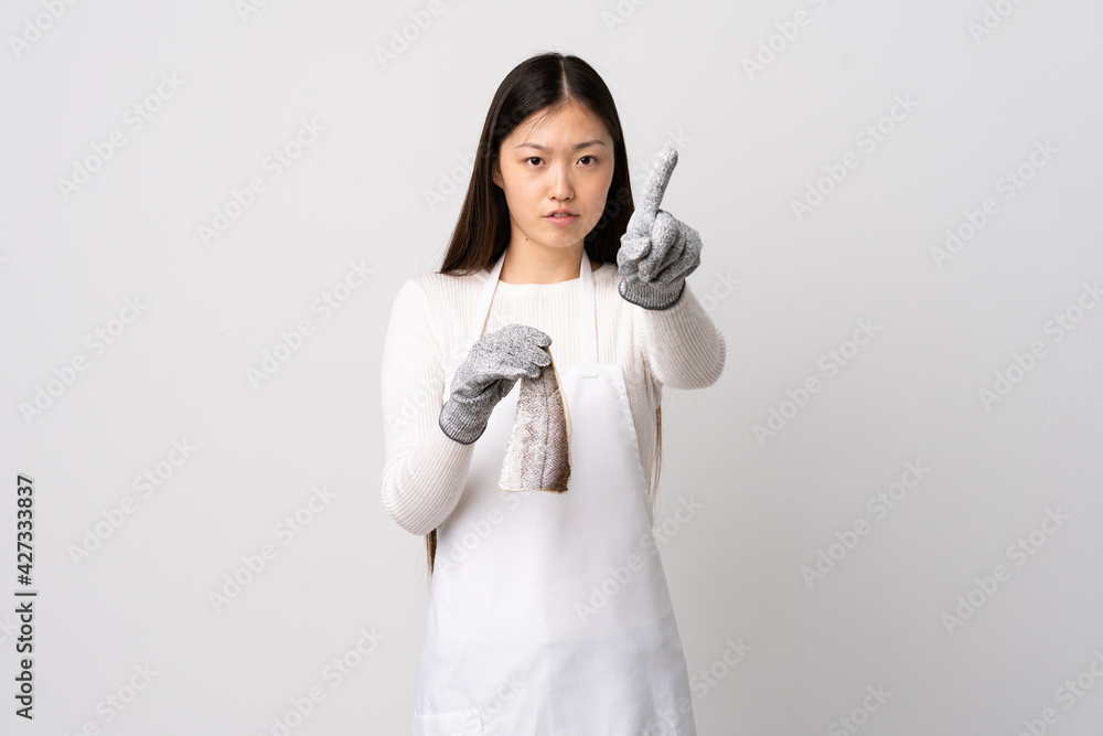 Chinese fishmonger wearing an apron and holding a raw fish over isolated white background counting one with serious expression Chinese fishmonger wearing an apron and holding a raw fish over isolated white background counting one with serious expression