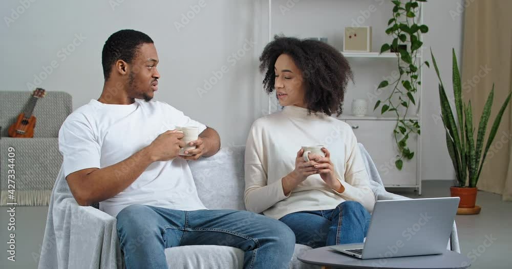 Couple afro american family young people husband and wife sitting on couch in cozy living room drinking hot tea coffee cocoa enjoying weekend together discussing what movie to watch online in laptop Couple afro american family young people husband and wife sitting on couch in cozy living room drinking hot tea coffee cocoa enjoying weekend together discussing what movie to watch online in laptop