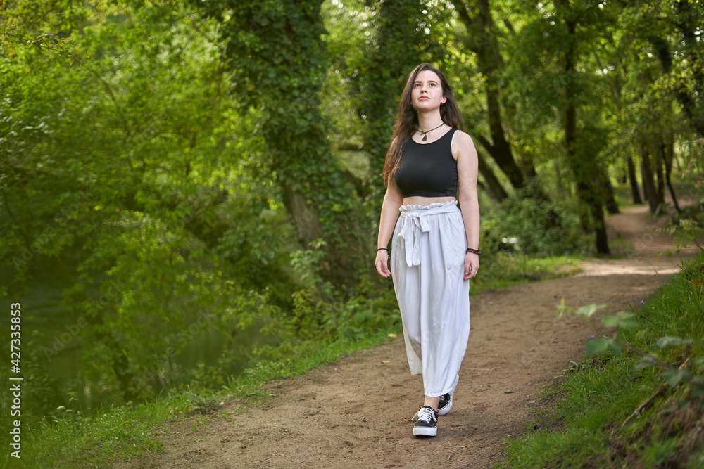 Young girl with dark hair and light skin in a black top, slouchy white pants, black and white sneakers and necklace `walking along a dirt path in lush green forest. woman in nature. teenager hiking Young girl with dark hair and light skin in a black top, slouchy white pants, black and white sneakers and necklace `walking along a dirt path in lush green forest. woman in nature. teenager hiking