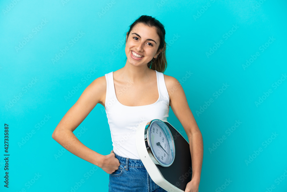 Young caucasian woman isolated on blue background with arms at hip and holding weighing machine Young caucasian woman isolated on blue background with arms at hip and holding weighing machine