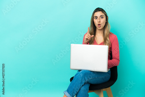 Young caucasian woman sitting on a chair with her pc isolated on blue background thinking an idea pointing the finger up © luismolinero