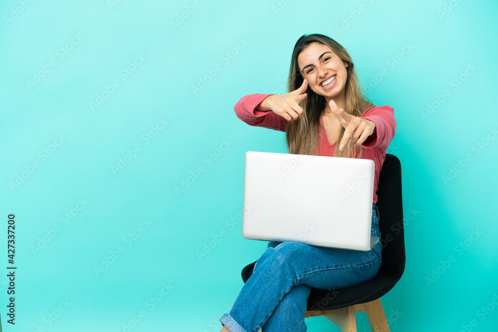 Young caucasian woman sitting on a chair with her pc isolated on blue background pointing to the front and smiling Young caucasian woman sitting on a chair with her pc isolated on blue background pointing to the front and smiling