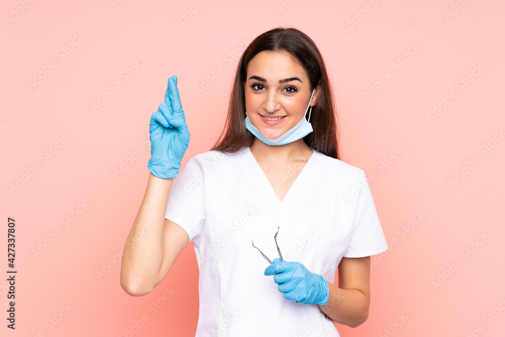 Woman dentist holding tools isolated on pink background with fingers crossing