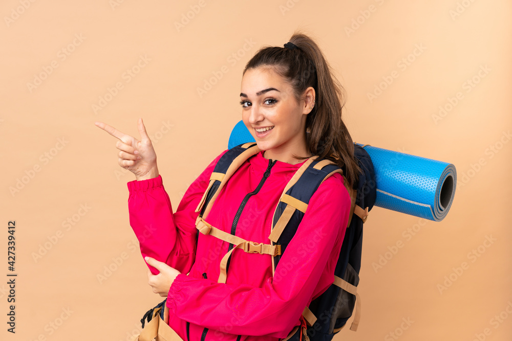 Young mountaineer girl with a big backpacker isolated on beige background pointing finger to the side Young mountaineer girl with a big backpacker isolated on beige background pointing finger to the side