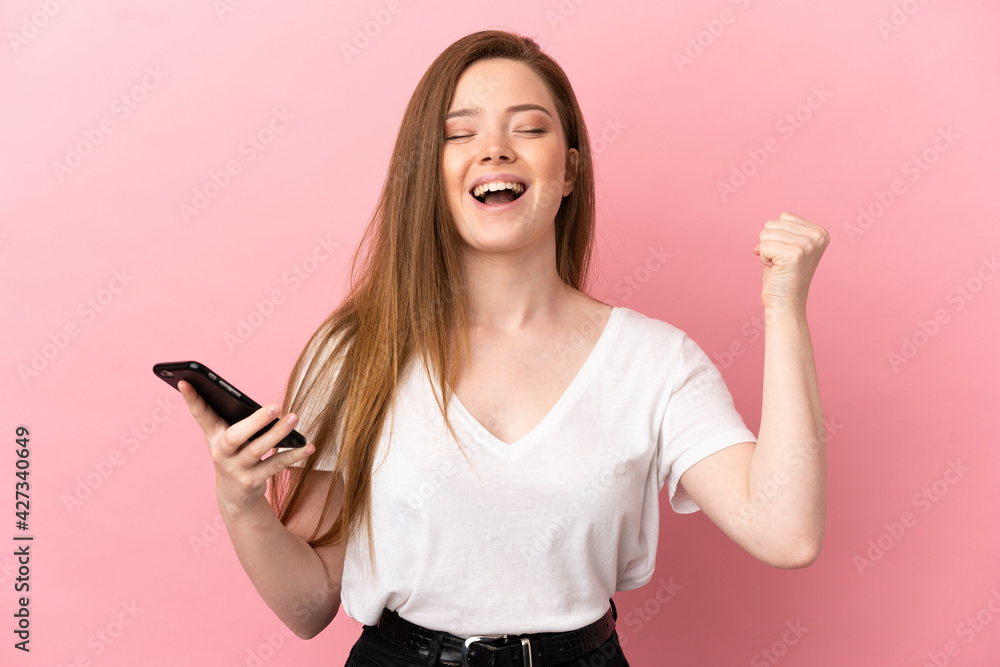 Teenager girl over isolated pink background using mobile phone and doing victory gesture Teenager girl over isolated pink background using mobile phone and doing victory gesture
