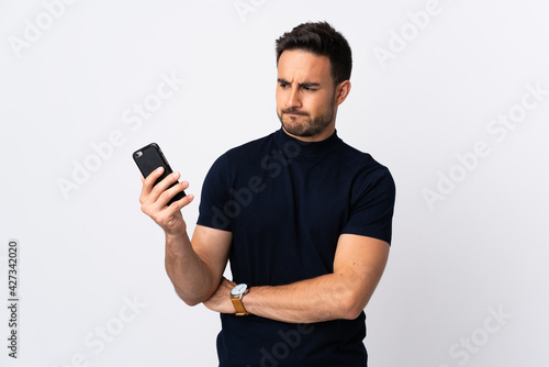 Young caucasian man using mobile phone isolated on white background with sad expression
