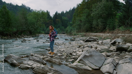 Woman standing at bank of river in forest. Girl hiking along mountain river