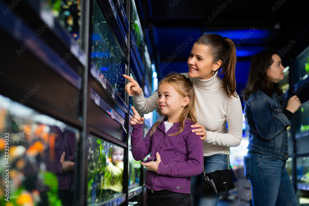Happy little girl with mother choosing new aquarium fish for home fish tank in pet store