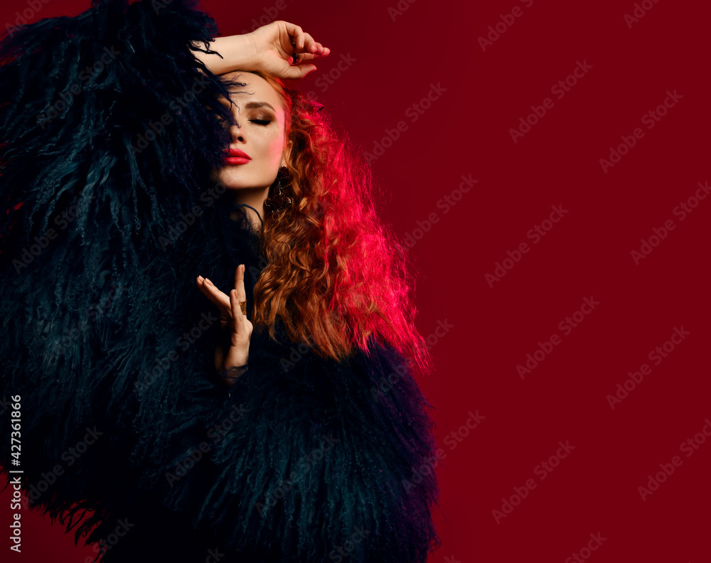Gorgeous rich young sensual woman with pouty lips, evening makeup and red curly hair wearing luxury fur coat is posing with eyes closed, arm at head isolated over red background