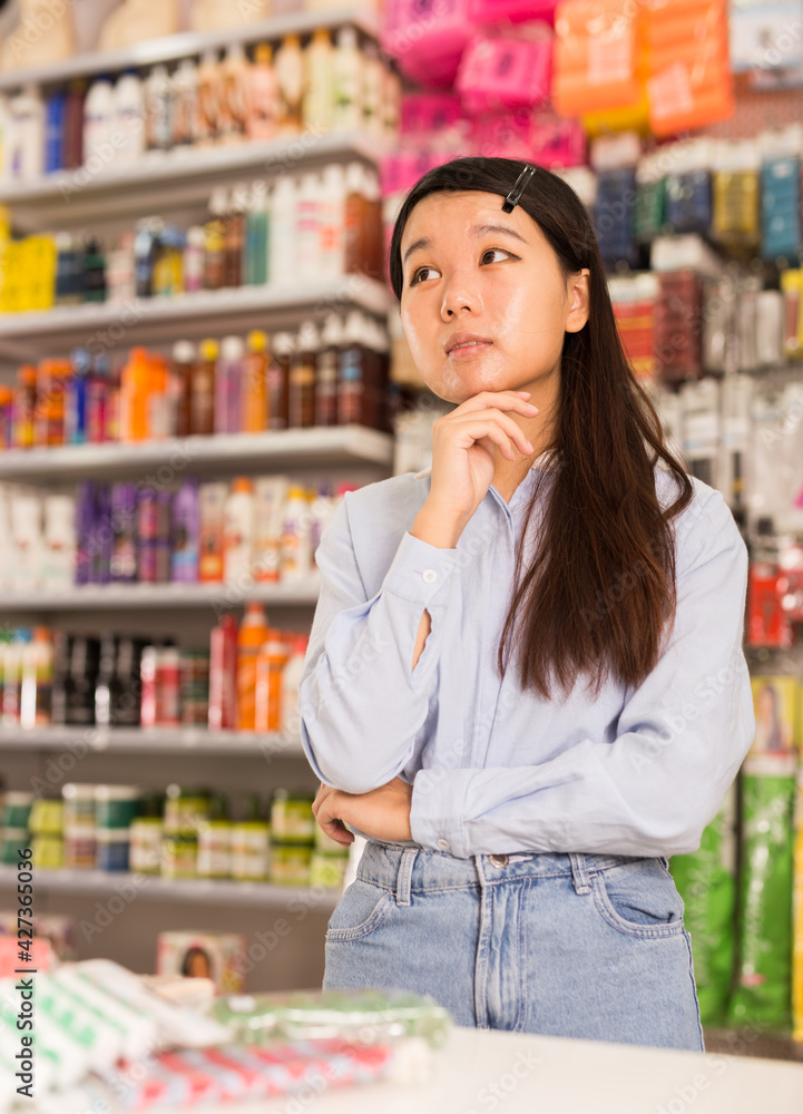 Portrait of puzzled Asian girl having doubts about choice of hair care products in special store