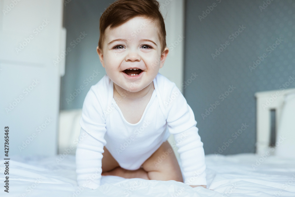 A portrait of a cute toddler baby boy in a white bodyuit seating on bed and laughing.Happy baby face look at the camera, in bedroom at home
