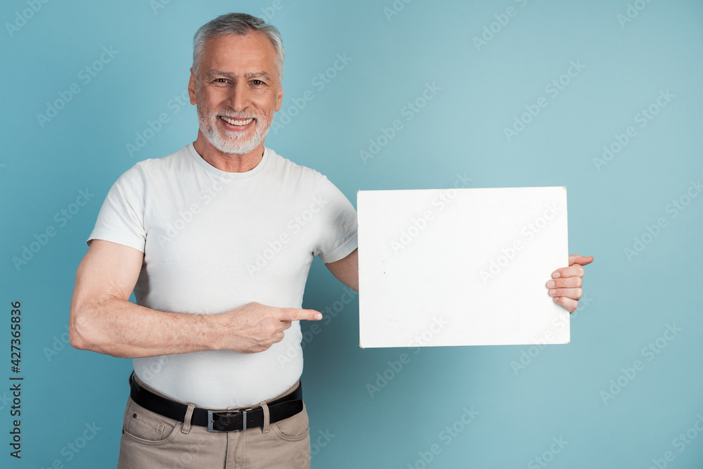 Smiling, pleasant, senior man holds a white piece of paper, points to it. Isolated on blue background man holding blank white piece of paper, copy space, place for advertising. Smiling, pleasant, senior man holds a white piece of paper, points to it. Isolated on blue background man holding blank white piece of paper, copy space, place for advertising.