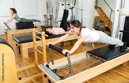 Positive young Latina practicing pilates stretching exercises on reformer at gym..