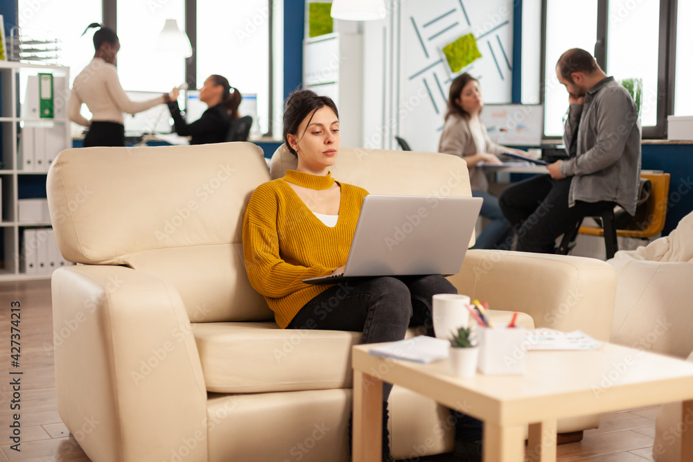 Fototapeta premium Succesfull business woman working on laptop on start up project sitting on cozy couch. Multiethnic professional company people coworkers discussing task in modern workplace.