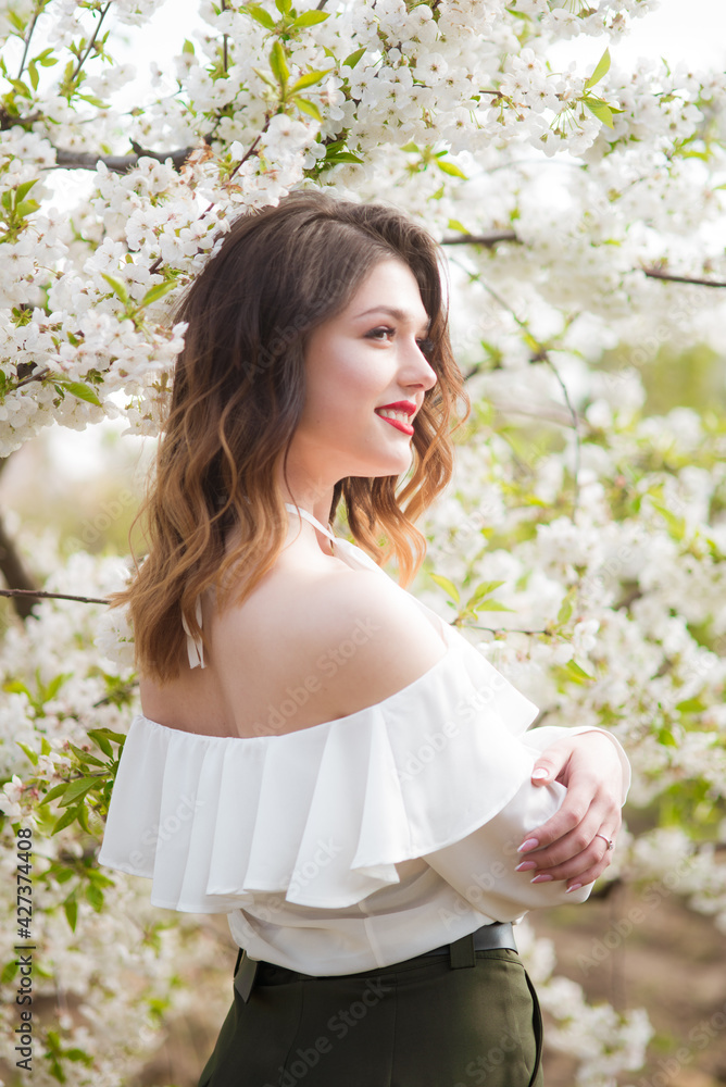 Lovely young girl in a white romantic silk blouse in spring in a blooming cherry garden in sunny weather. Spring and Fashion. Spring blooming gardens Lovely young girl in a white romantic silk blouse in spring in a blooming cherry garden in sunny weather. Spring and Fashion. Spring blooming gardens