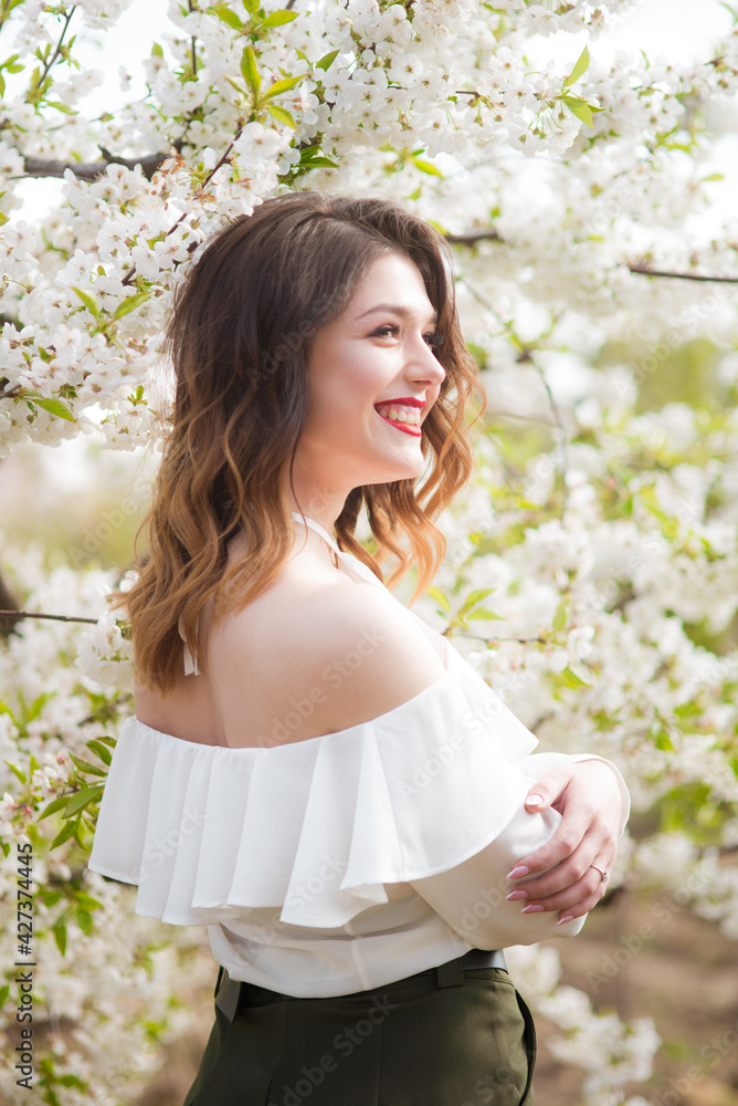 Lovely young girl in a white romantic silk blouse in spring in a blooming cherry garden in sunny weather. Spring and Fashion. Spring blooming gardens