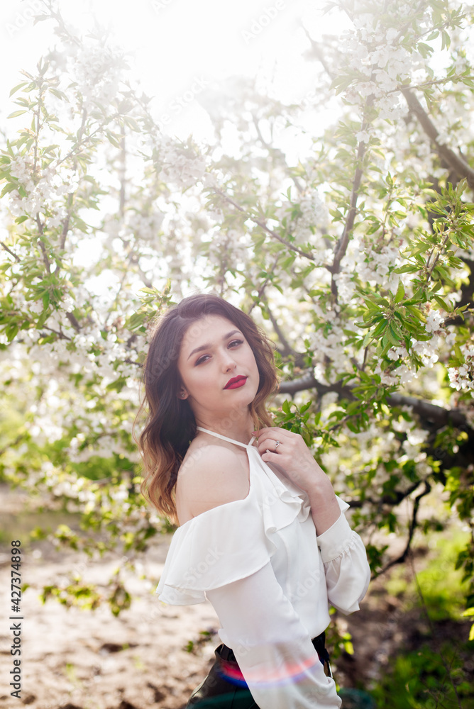 Lovely young girl in a white romantic silk blouse in spring in a blooming cherry garden in sunny weather. Spring and Fashion. Spring blooming gardens Lovely young girl in a white romantic silk blouse in spring in a blooming cherry garden in sunny weather. Spring and Fashion. Spring blooming gardens