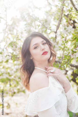 Lovely young girl in a white romantic silk blouse in spring in a blooming cherry garden in sunny weather. Spring and Fashion. Spring blooming gardens