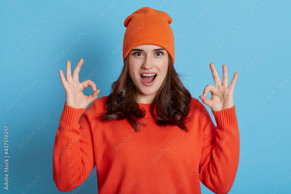 Young European excited female wearing orange sweater and cap looking at camera with opened mouth and showing ok signs with both hands, dark haired girl isolated over blue background. Young European excited female wearing orange sweater and cap looking at camera with opened mouth and showing ok signs with both hands, dark haired girl isolated over blue background.