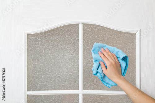 Young adult female hand holding blue dry rag and wiping glass of white wooden door in room. Closeup. Front view. General or regular cleanup. Cleaning service. photo