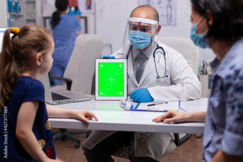 Doctor with face mask and visor using green screen tablet pc in examination office during coronavirus pandemic. Healthcare physician with chroma key notebook isolated mockup screen