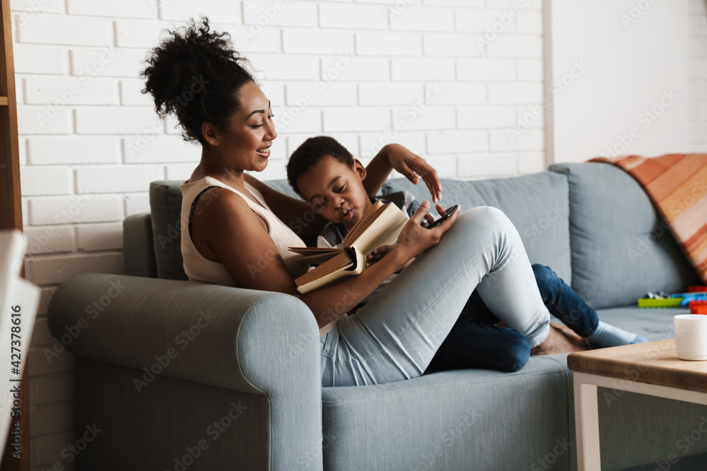 Obraz premium Black pleased mother and son reading book while sitting on sofa at home