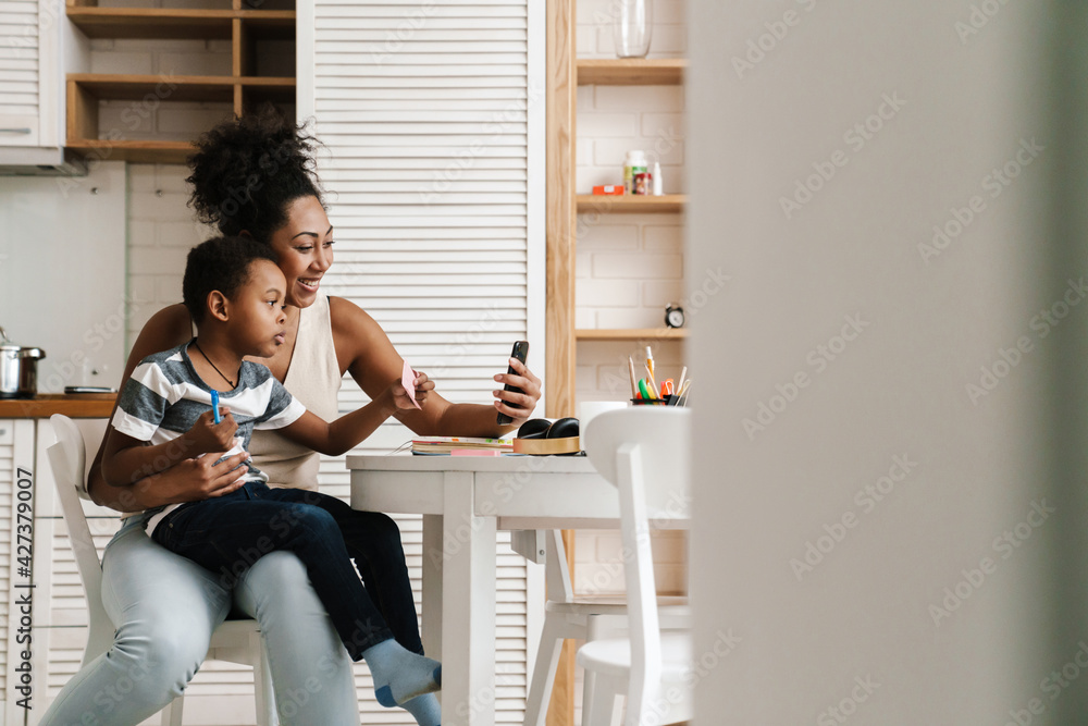 Black smiling mother and son using mobile phone while sitting together Black smiling mother and son using mobile phone while sitting together