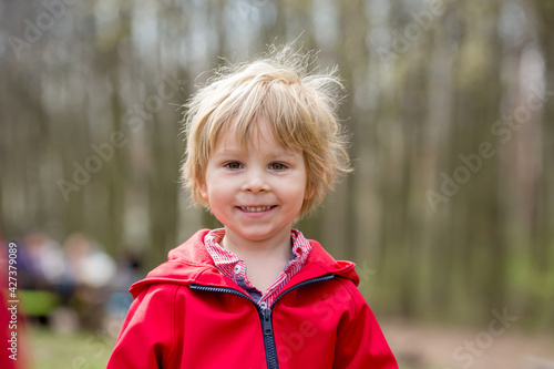 Close portrait of smiling toddler blond child, cute boy with red jacket in the park