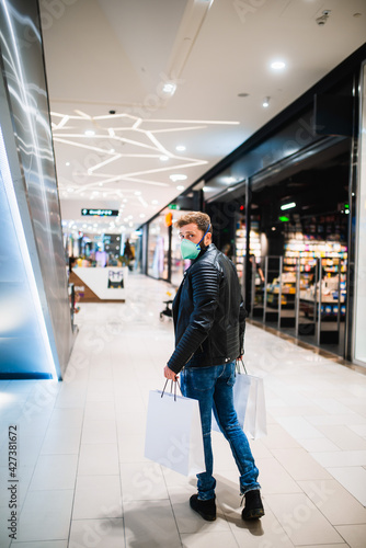 A young bearded caucasian man in a black leather jacket, with a mask and bags in his hand walks in the mall during the epidemic COVID - 19 coronavirus