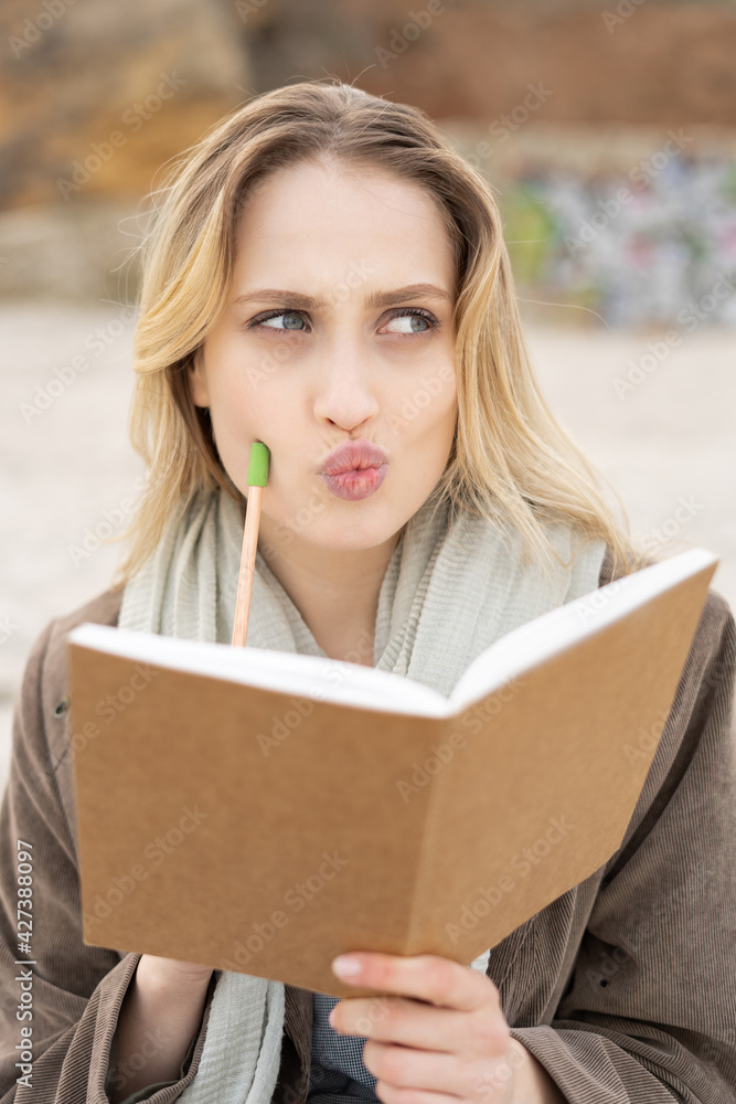 Portrait of a charming young girl thinking and holding a notepad with a pen on her chick looking aside on a beach Portrait of a charming young girl thinking and holding a notepad with a pen on her chick looking aside on a beach