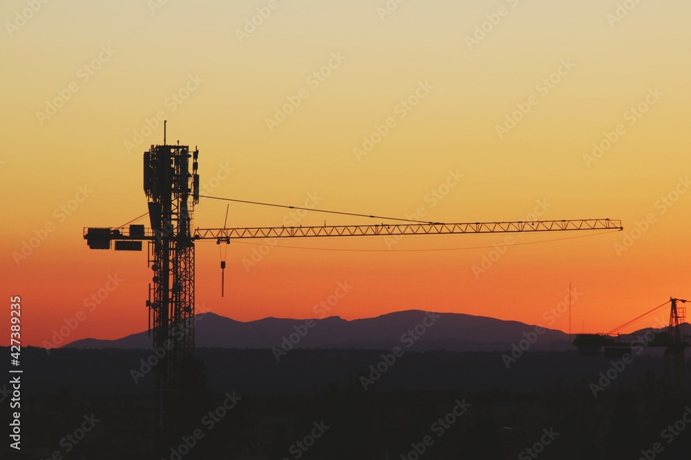 Fototapeta premium Grúa torre de construcción al atardecer. Silueta de la grúa sobre un bonito fondo anaranjado con los últimos rayos de sol en Madrid, España.