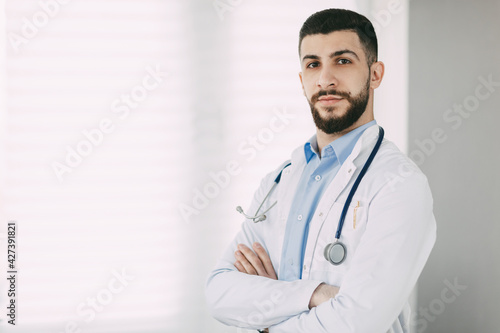 Serious young doctor in stethoscope and white medical coat stands in his office with arms crossed