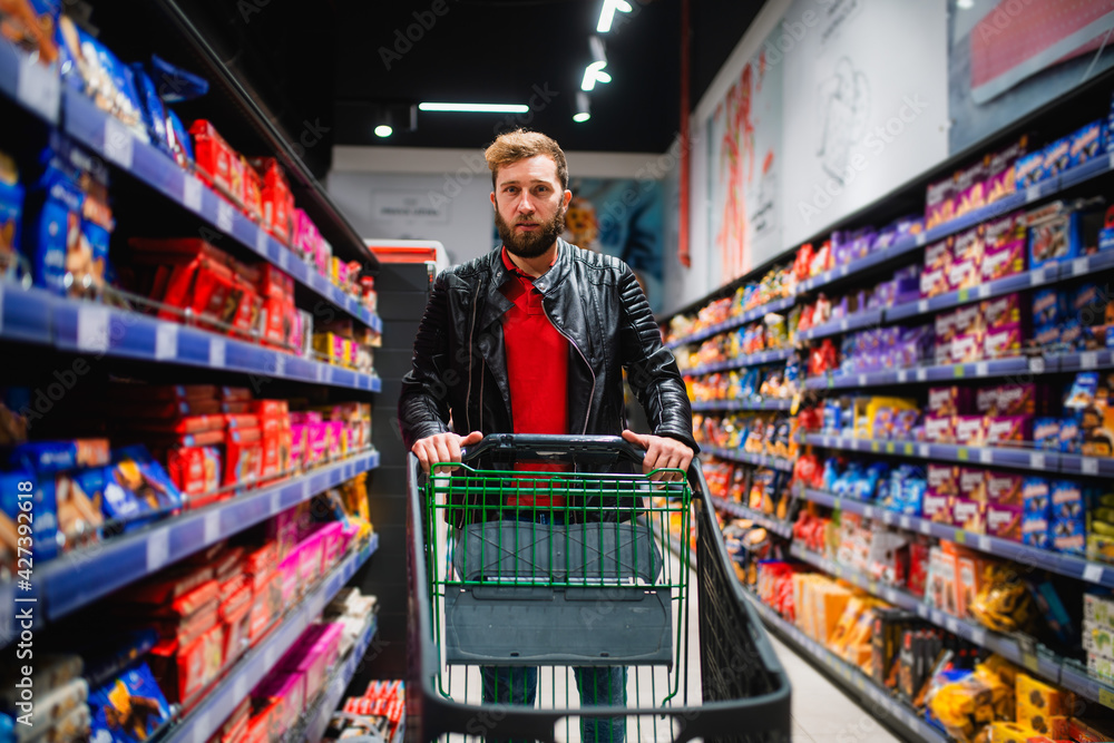 A young handsome bearded man buys groceries at the supermarket and pushes a basket of food