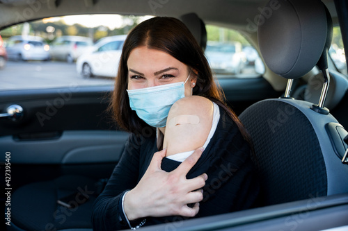 Portrait of a young woman in car shows arm with patch, smiling after having the vaccine against Coronavirus Covid-19 immunizing in the forecourt of the hospital - Person bares shoulder after injection