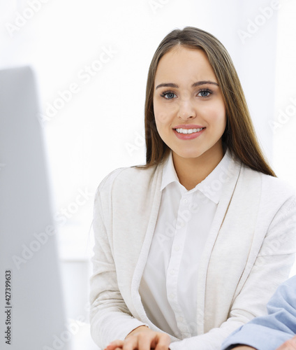 Young cheerful woman sitting at the desk with computer and looking at camera in white colored office. Looks like student girl or business lady. Headshot