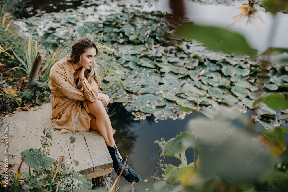 Fototapeta premium Young woman wearing a maxi chiffon dress sitting on a wooden pier by a pond with water lilies.