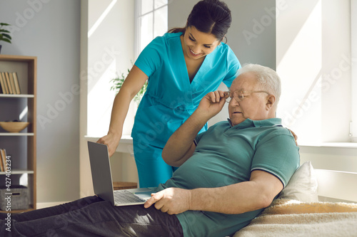 Nursing home, caregiving and elderly people assistance service. Friendly smiling nurse showing good analysis result or help with online call on laptop to old senior man patient lying on hospital bed photo