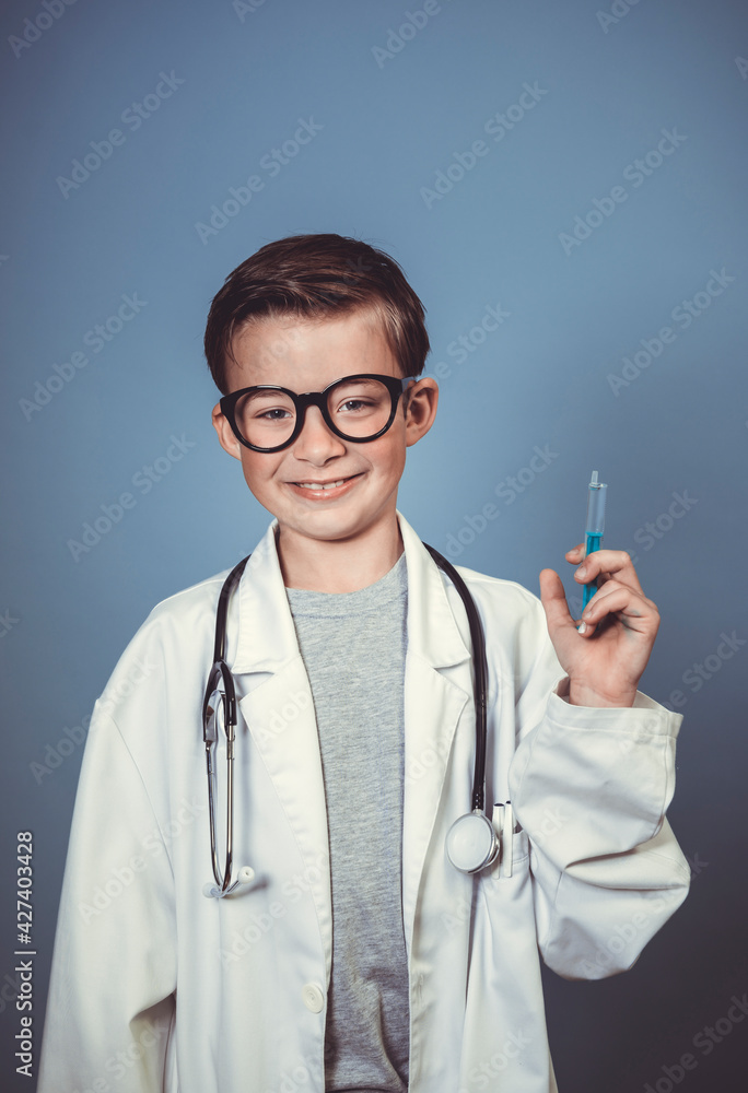 cool young boy is disguised as a doctor with a white doctor smock and plays with syringe and stethoscope in front of blue background in the studio cool young boy is disguised as a doctor with a white doctor smock and plays with syringe and stethoscope in front of blue background in the studio