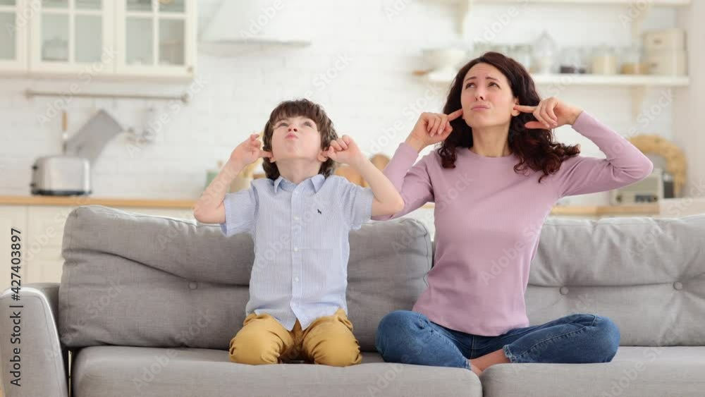 Frowning mom and son sit on couch with closed eyes and cover ears from noisy music or fight sounds from neighbors. Young female parent and kid tired of drilling noise not listen plug ears with fingers Frowning mom and son sit on couch with closed eyes and cover ears from noisy music or fight sounds from neighbors. Young female parent and kid tired of drilling noise not listen plug ears with fingers